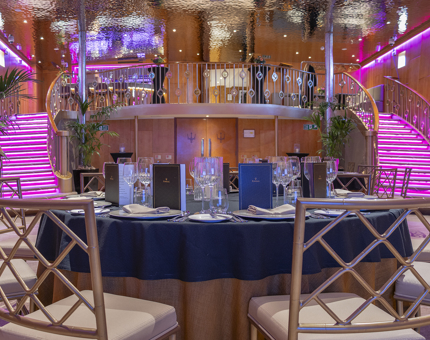 The double height Ballroom of Fingal Hotel. In the foreground there is a table set for dinner. There are two curved staircases in the background leading to a bar area. There is pink lighting 