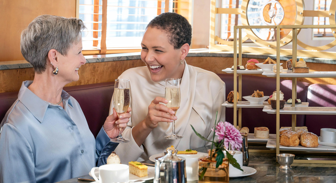 Two women holding champagne glasses sitting at a table with two afternoon tea stands on it. 