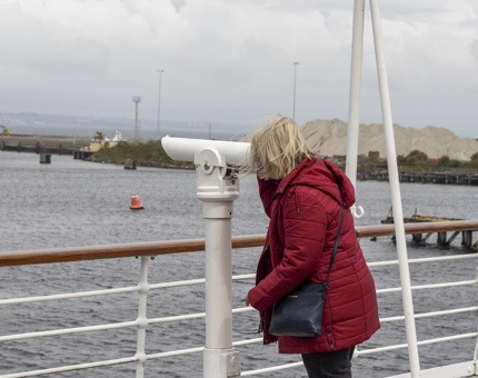 A woman wearing a red jacket looking through a telescope on the Verandah Deck. There is a view of the docks in the background. 