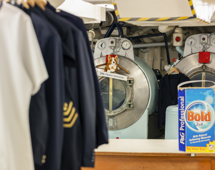 Britannia's onboard Laundry. There are sailor's uniforms hanging in the foreground and industrial sized washing machines in the background. 