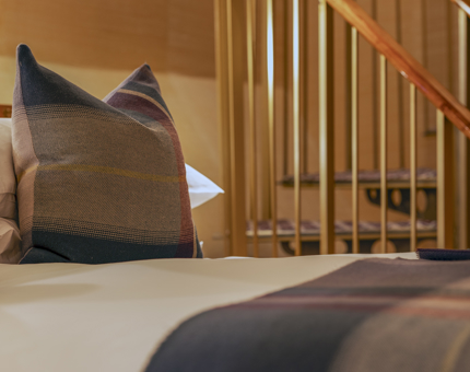 A close up of a bed with white sheets and a tartan blanket and pillow. There is a spiral staircase in the background. 