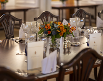 A dining table set with menus at each place. There are orange flowers in a vase on the table. 