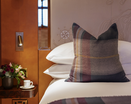 A bed with white sheets and a tartan pillow and blanket. There is a side table with a plant and white tea cup and saucer on it.