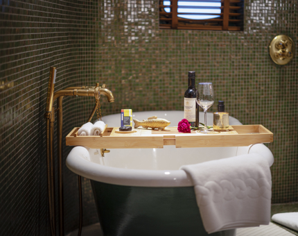 A roll-top bath with a wooden tray over it. There is a bottle of wine, a bar of chocolate, and a flower on it.