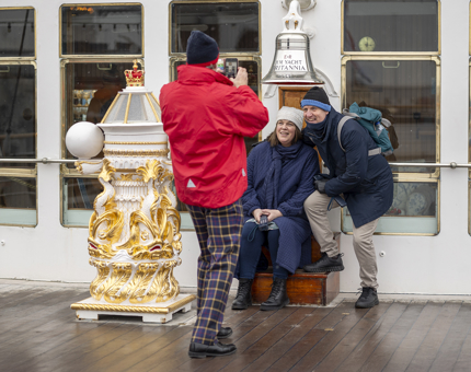 A visitor assistant wearing a red jacket is taking a photo of two visitors at Britannia's Bell. 
