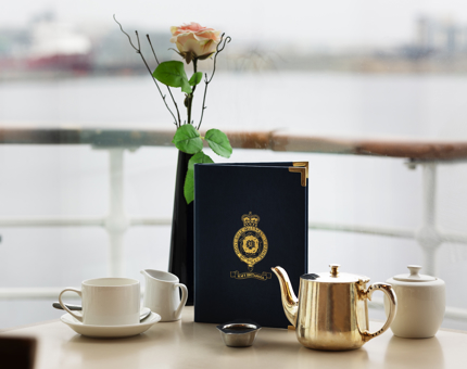 A table in the Tearoom set with a tea pot, milk jug, menu and vase with a flower in it. There is a view of the waterfront out of the window behind the table.
