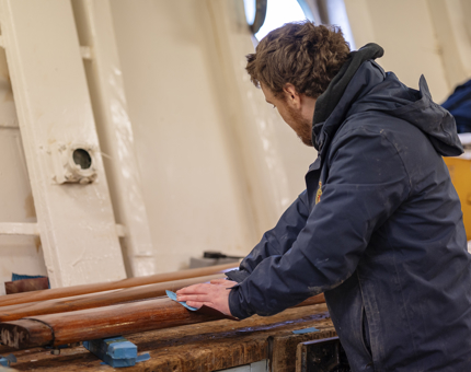 A Maintenance team member wearing a blue jacket sanding wooden handrails.