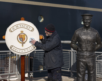 A facilities Officer wearing a navy blue jacket and hat is wiping the Perry Bout cover on the quayside. Next to him is a statue of an ex-Royal Yachtsman, Norrie. Britannia is in the background. 