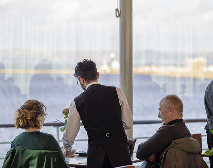 A member of the Tearoom waiting team is serving two visitors at their table. The table is situated in front of large picture windows with views out over the port. 