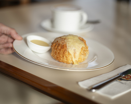 A hand is holding a white plate with a cheese scone and pot of butter on it. It is being placed on a table. 
