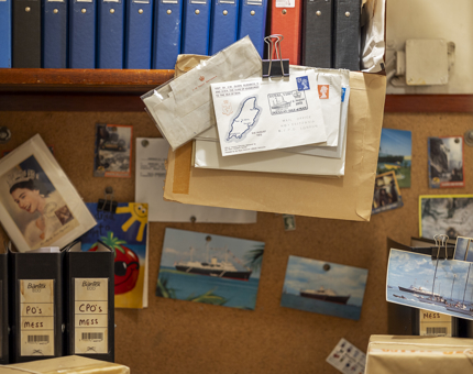 The onboard Post Office. There is a pile of letters hanging from the shelf by a bull dog clip. There are lots of ring binders lined up on the shelf. On the corkboard in the background, there are many post cards pinned to it. 
