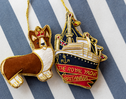 A corgi and Britannia ship hanging decorations. They are sitting on a blue and white stripped background. 