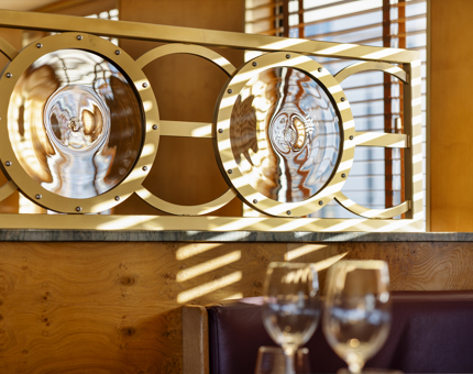 A close up of the round nautical glass and brass details in The Lighthouse Restaurant aboard Fingal Hotel. There is sun streaming in from the windows from the right hand side. In the foreground, there are two wine glasses on the table. 