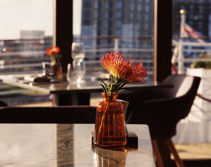 A close up of a an orange flower in an orange vase on a table. There is another table in the background with the same flower on it. 