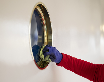 An arm wearing a red fleece and hand wearing a blue glove is holding a cloth, wiping a porthole.