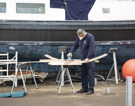 A Maintenance team member is outside next to a boat, he is sanding a frame for the boat.