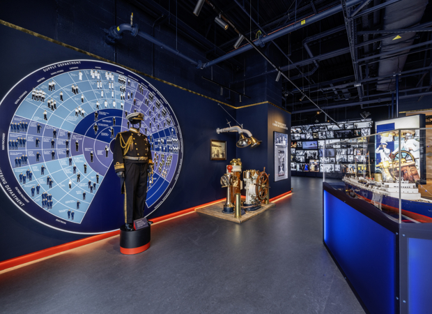 Inside the brand new Visitor Centre at Britannia. The Admiral's uniform is displayed in front of a wall display of a wheel that details all of the different job roles aboard. To the right is a model of Britannia and in the background is a cascading wall of images and video. 