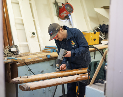 A man is in the Maintenance workshop using a heatgun to remove varnish from a wooden handrail.