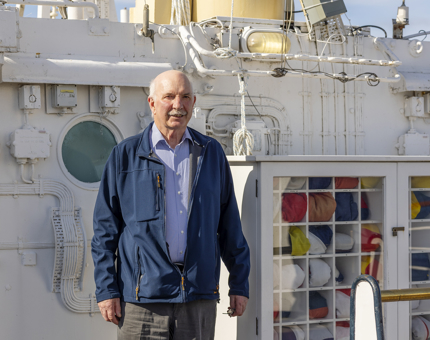 A man standing on the Bridge of Britannia next to the flag locker which is filled with folded colourful flags. 
