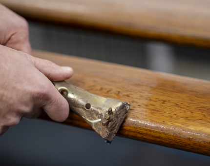 A close up of a hand holding a scraper, taking the varnish off a handrail.