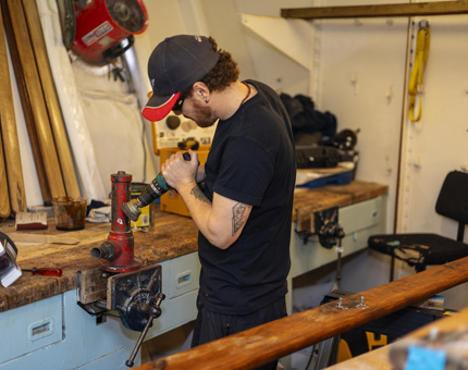 In the workshop paint is being removed from a seawater strainer from the Fast Motor Launch boat. The Maintenance team member is standing at a bench with his back towards the camera. 