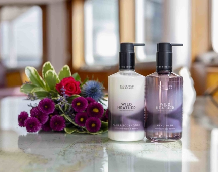 Two bottles of Wild Heather bath and body products on a table, there is a bunch of flowers next to them. 