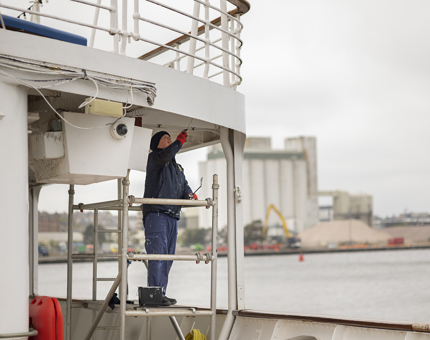 On the starboard side quarter deck, a Maintenance man is standing on some scaffolding holding a paintroller, applying a coat of paint.