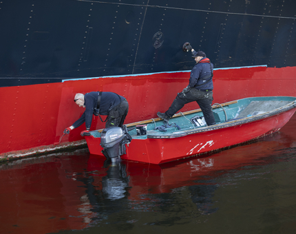 Two Maintenance team members on a small boat alongside Fingal. One is painting with red paint, the other is holding the boat steady.