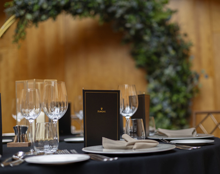 In Fingal's Ballroom, there is a table set for dinner. It has a dark blue tablecloth and had wine glasses, plates and cutlery on it. In the background there is an arch made from green foliage.
