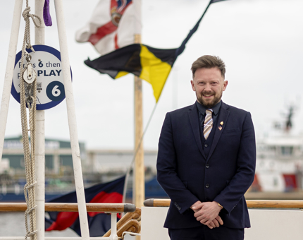Matt, HR Coordinator, posing for a photo on the Verandah Deck. There are dress flags behind him.