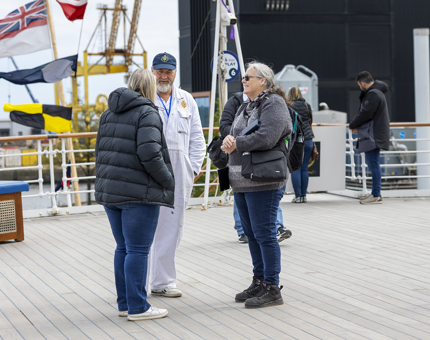 On the Verandah Deck, a Yottie is speaking with two visitors. There are flags flying in the background. 