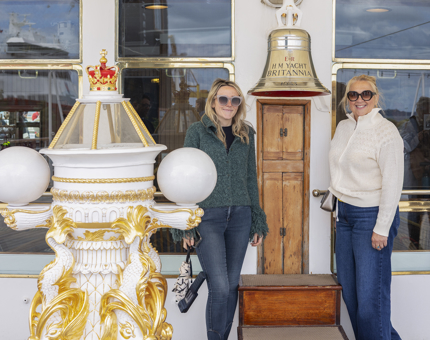 Two women standing next to The Royal Yacht Britannia's Bell and binnacle on the Verandah Deck. 