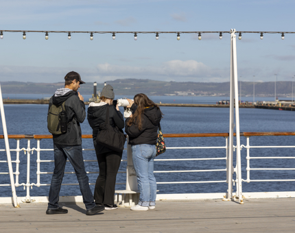 Three visitors on the Verandah Deck, one is looking through the telescope. There are views of the water and hills in the background. 