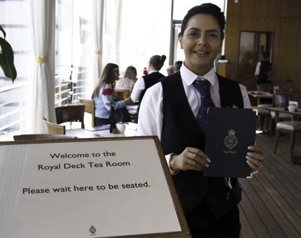Woman holds Britannia menu and waits to greet visitors to the Tea Room. 