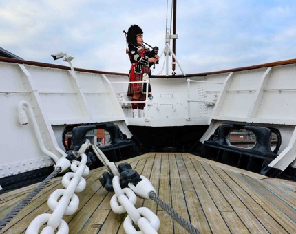 Piper plays the bagpipes on Britannia's deck while Union Jack flies at half mast. 