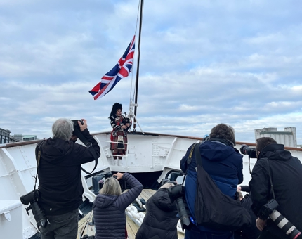 Photographers take pictures of a piper playing the bagpipes on deck. 