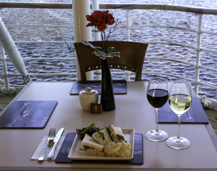 Table laid for lunch with a plate of sandwiches, salad and crisps and two glasses of red and white wine with a sea view in the background. 