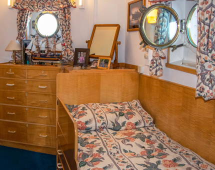 Inside The Admiral's Sleeping Cabin with sycamore veneered bed and chest of drawers, floral curtains and family photographs on a cabinet. 
