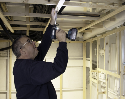 Maintenance team member drills into wood in the ceiling in the Training Room. 