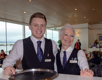 Two Waiters, a man and a woman, pose smiling for a photo in the Royal Deck Tearoom. 