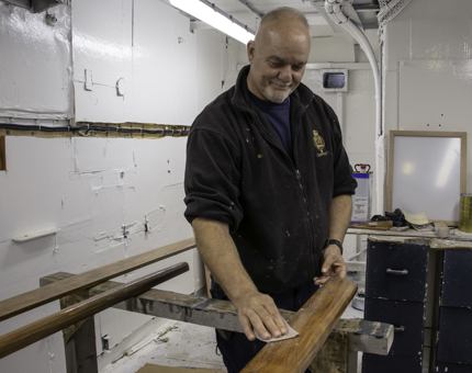 A man from the Maintenance team is sanding wooden handrails in the workshop. 