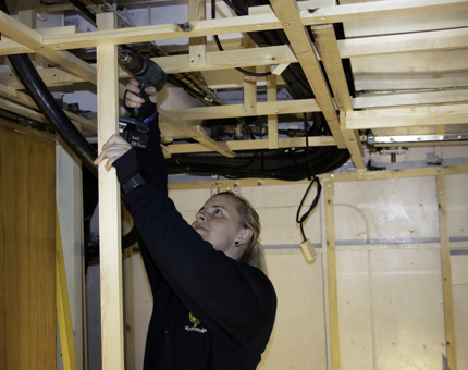 A woman from the Maintenance team is using a drill to secure a wood support into position. 