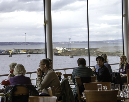 Several visitors sit at tables in The Royal Deck Tea Room overlooking a sea view. 