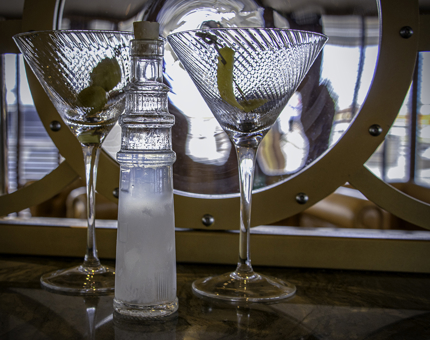 Two martini glasses, one containing olives and one containing a twist of lemon alongside a lighthouse-shaped bottle. 