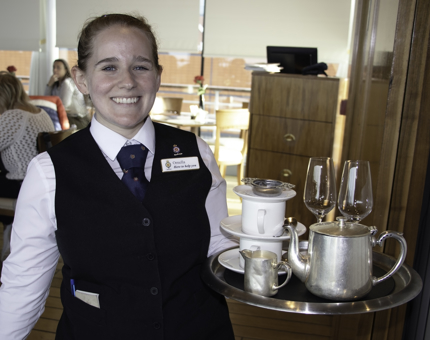A smiling waiter holds a tray containing a teapot, cups, saucers and glasses.
