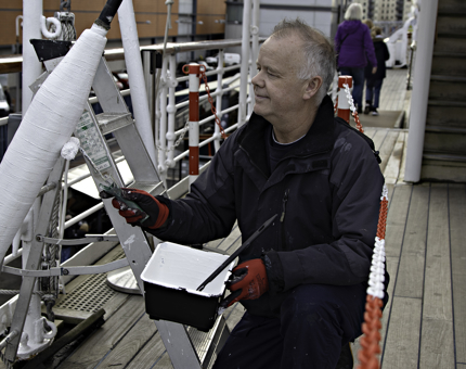 A man from Maintenance is using a roller to paint parts of the Verandah Deck with white paint. 