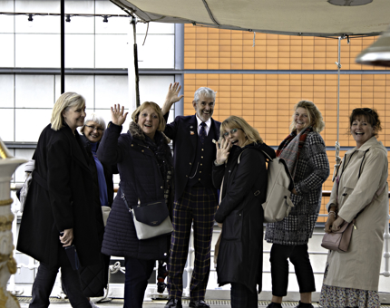 Man Visitor Assistant stands with a group of women as they all wave to the camera. 