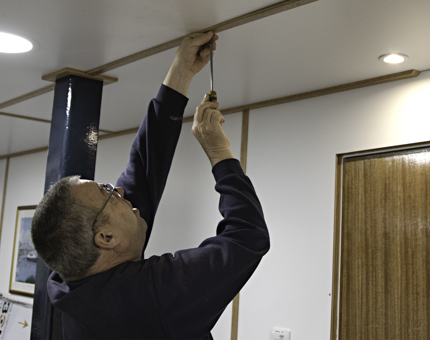 Man uses a screwdriver to affix beading to the ceiling in the Junior Rates Dining Hall.