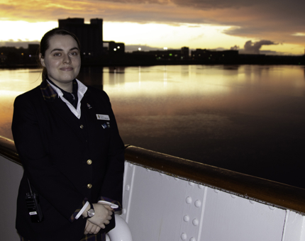 Woman Visitor Assistant stands on Britannia's outside deck in front of a vibrantly coloured sunset. 