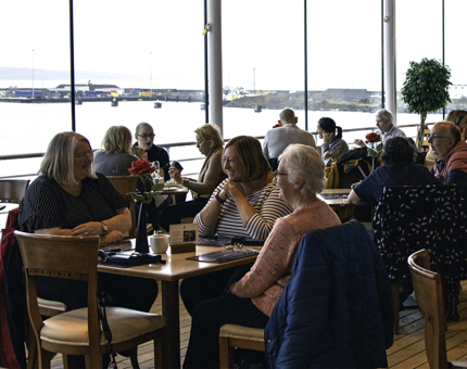 Small groups of visitors sit at tables enjoying food and drinks in the Royal Deck Tea Room. 